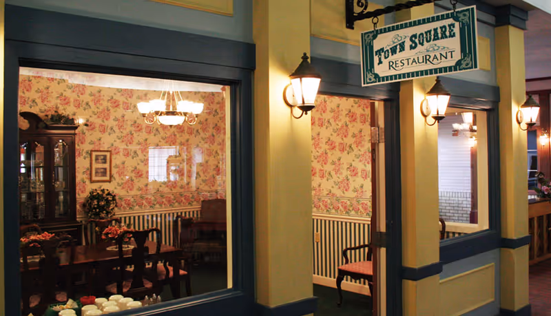 View through windows into a floral‑papered dining room labeled "Town Square Restaurant" with wooden tables, chairs, and chandeliers.