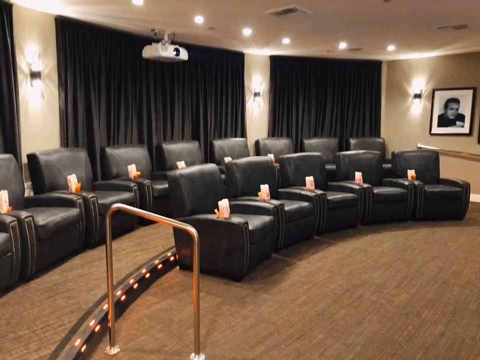 Small private theater room with curved rows of black leather recliners, a ceiling projector and dim wall lighting.