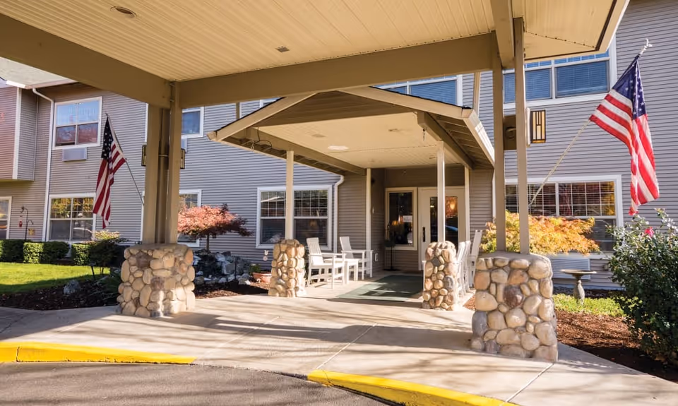 Covered entryway of a senior living building with stone column bases, white chairs, and two American flags.