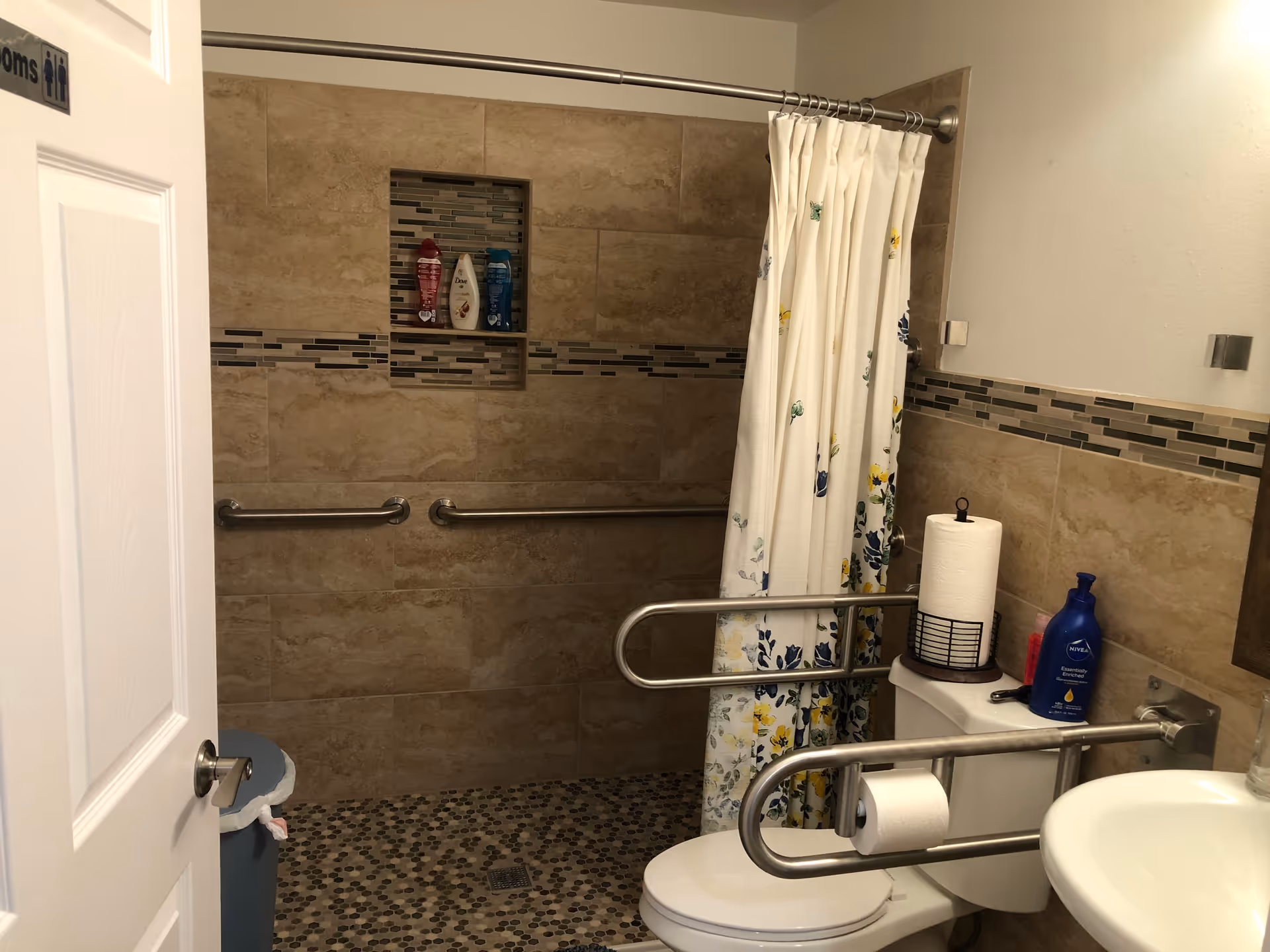 Accessible bathroom with tiled walk-in shower, floral shower curtain, grab bars, toilet with fold-down rails, and a sink.