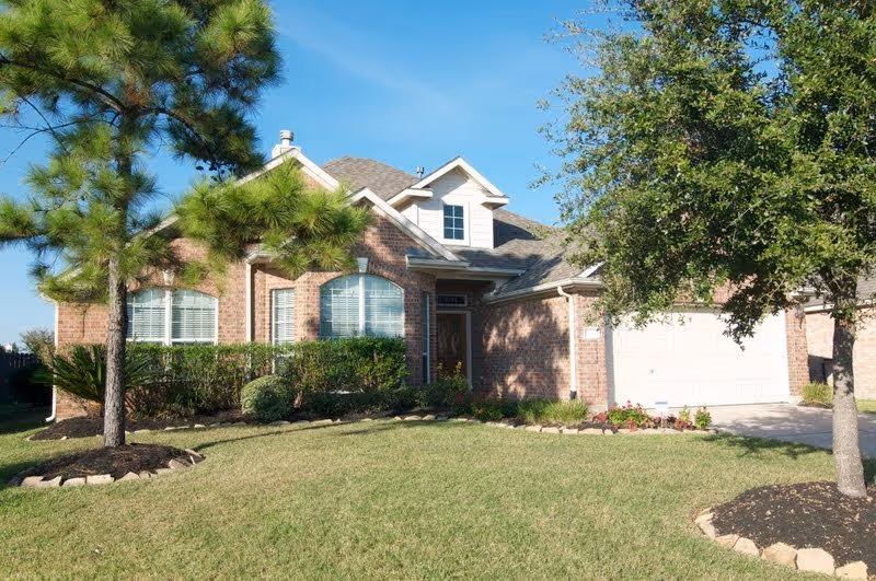A single-story brick house with a well-maintained front yard featuring green grass, two trees, and trimmed bushes. The house has a large window with white blinds, a front door with sidelights, and a two-car garage. The sky is clear and blue.