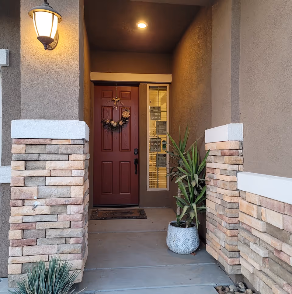 A front porch entrance with a red door decorated with a floral wreath. The porch has stone pillars with beige and light brown bricks, a potted plant on the right side, a doormat, and a lit wall-mounted lantern on the left.
