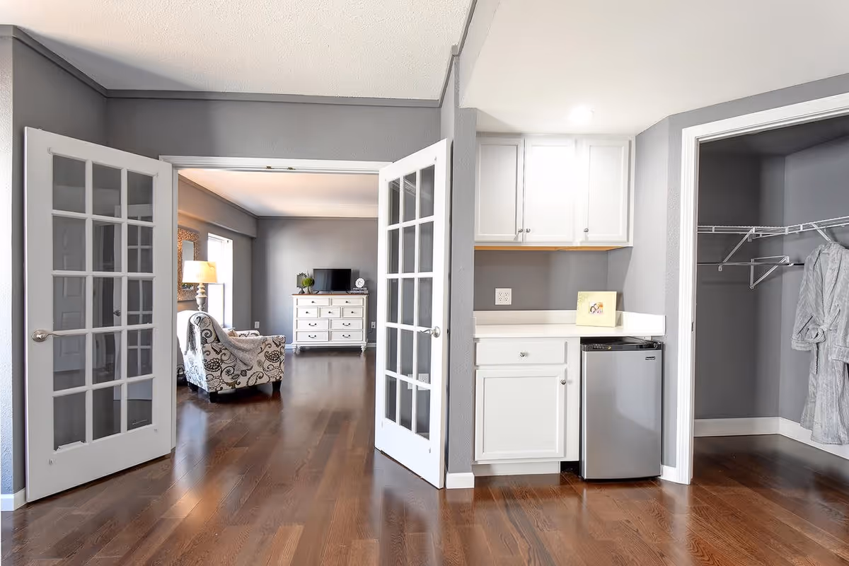 Interior view of a senior living facility showing an open space with wooden flooring. On the left, there are white French doors leading to a room with a patterned armchair, a lamp, and a dresser with a TV on top. On the right, there is a small kitchenette area with white cabinets, a countertop, and a mini refrigerator. Next to the kitchenette is an open closet with wire shelving and a hanging robe.