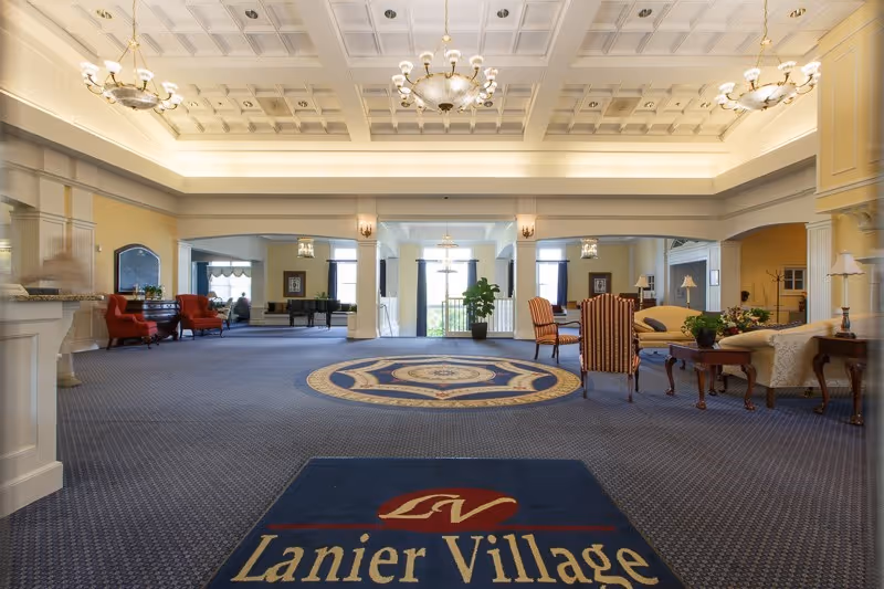 Spacious, well-lit lobby with seating areas, decorative coffered ceiling, and a floor mat reading "Lanier Village".