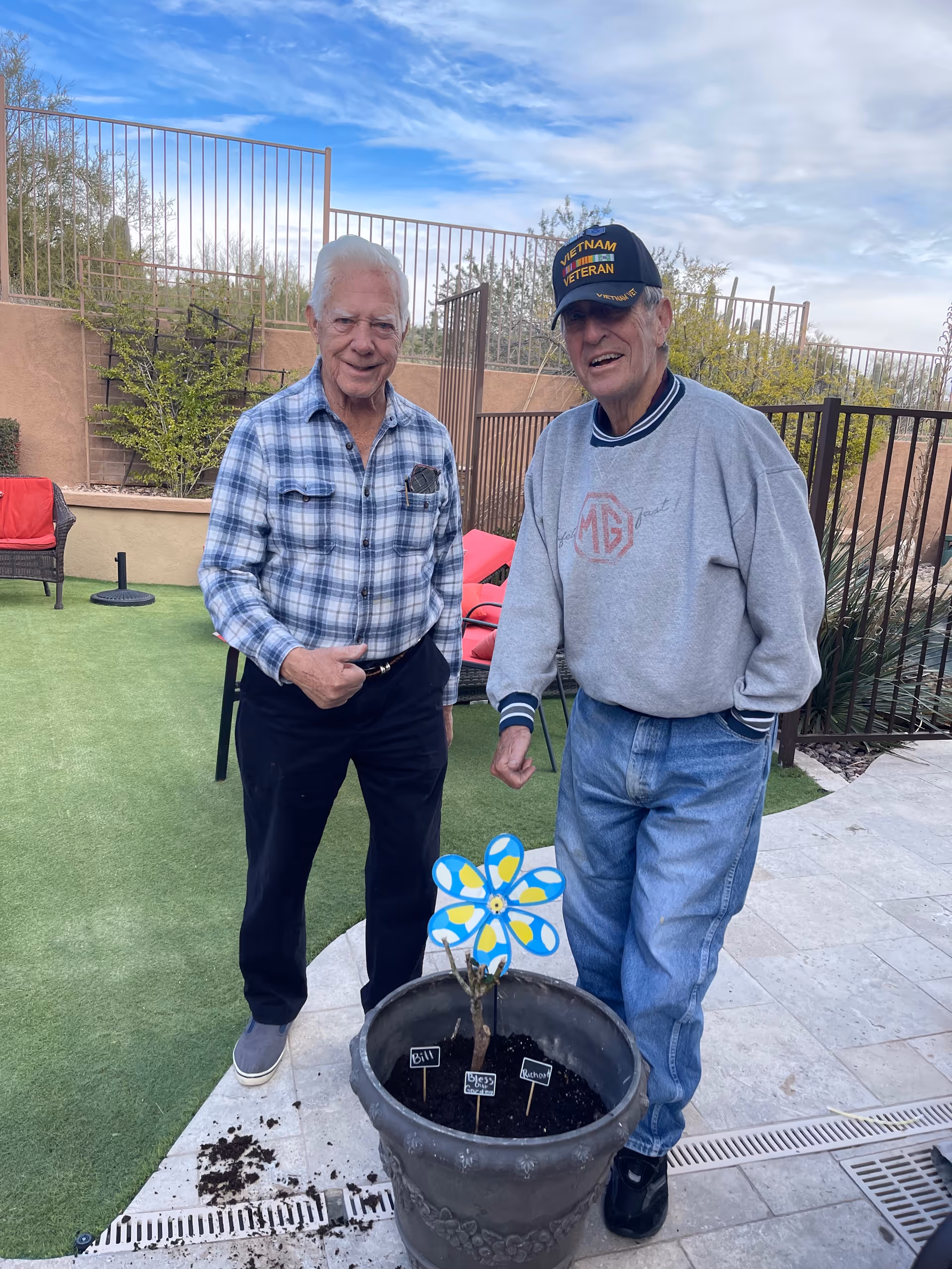 Two elderly men standing outdoors on a patio next to a large flower pot with soil and a decorative blue and yellow pinwheel. One man is wearing a plaid shirt and dark pants, the other is wearing a gray sweatshirt, jeans, and a Vietnam Veteran cap. Behind them is a fenced area with some greenery and outdoor furniture.