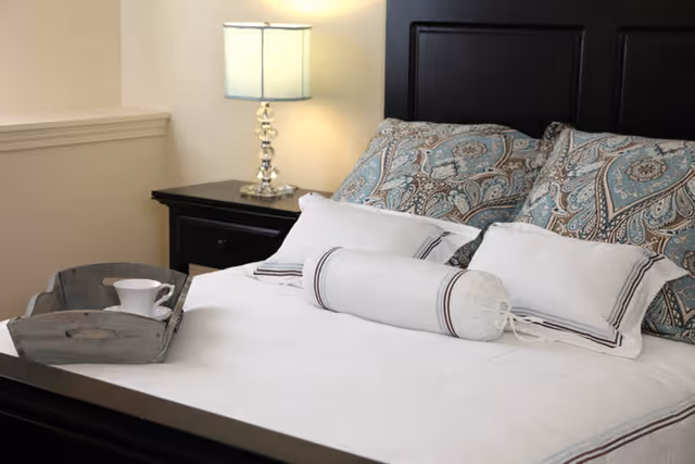 A neatly made bed with white bedding and decorative pillows featuring a blue and brown paisley pattern. A cylindrical bolster pillow is placed in front of two rectangular pillows. A wooden tray with a white cup and saucer sits on the bed. Next to the bed is a black nightstand with a clear glass lamp with a white lampshade. The wall behind the bed is light-colored.