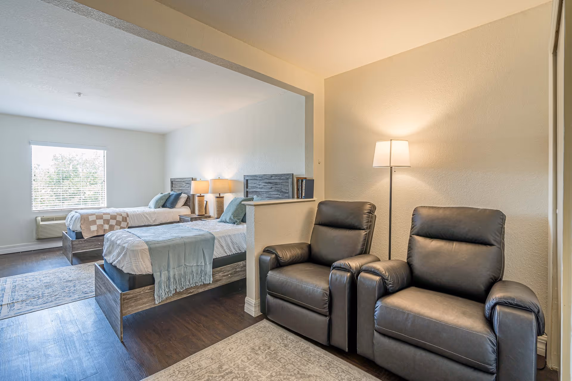 A bright and clean assisted living facility room featuring two twin beds with wooden headboards, bedside tables with lamps, and a window with blinds. In the foreground, there are two black leather recliner chairs next to a floor lamp, separated from the beds by a half wall.