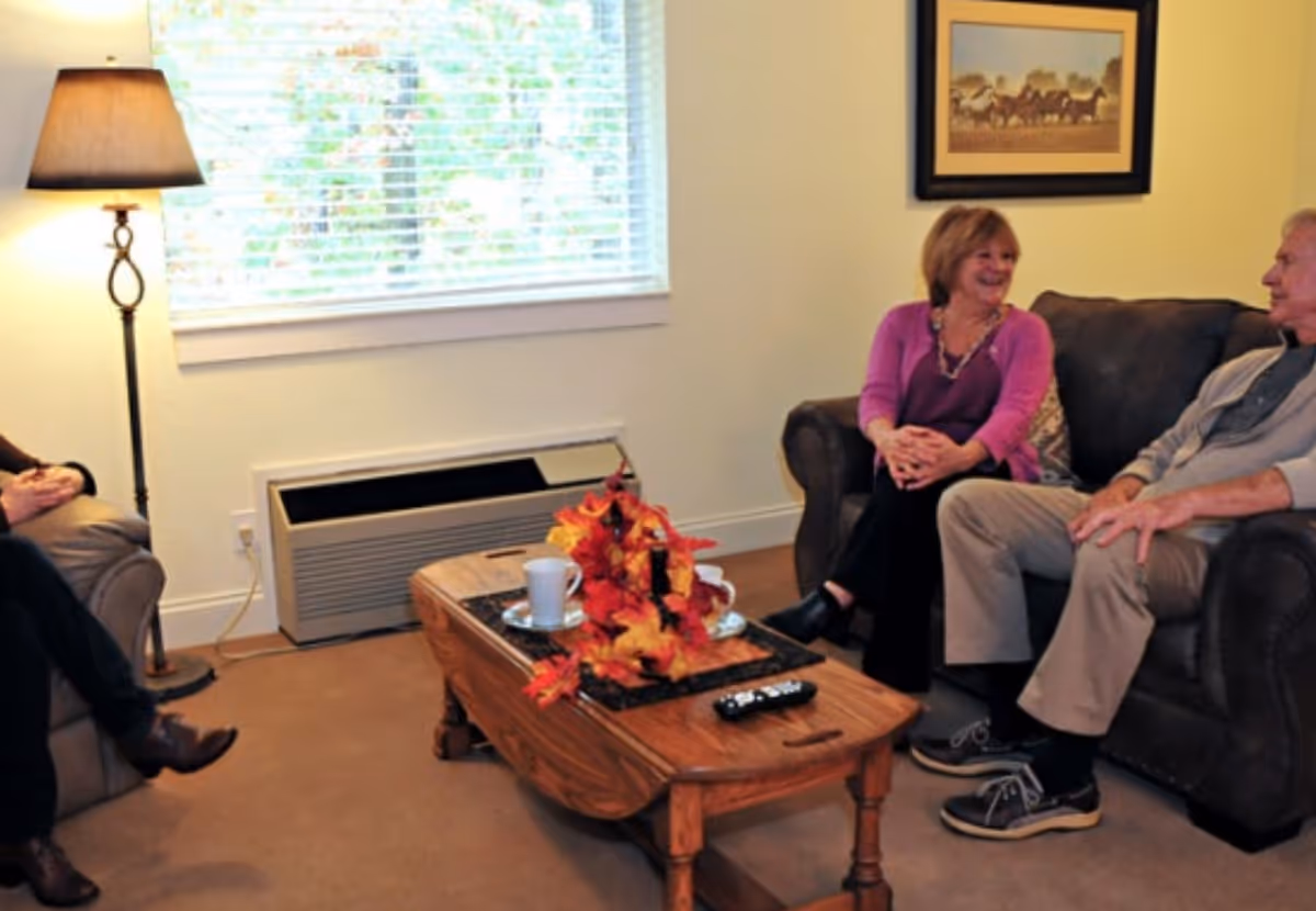 A cozy living room with three people sitting and chatting. There is a wooden coffee table in the center with a decorative autumn-themed centerpiece, a remote control, and a cup on a saucer. A floor lamp stands near a window with blinds, and a framed picture hangs on the wall behind the couch.