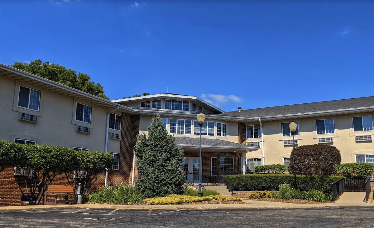 Exterior view of The Meadows of Franklin Grove Assisted Living facility showing a two-story building with multiple windows, a central entrance with a covered porch, surrounded by trimmed bushes and trees under a clear blue sky.