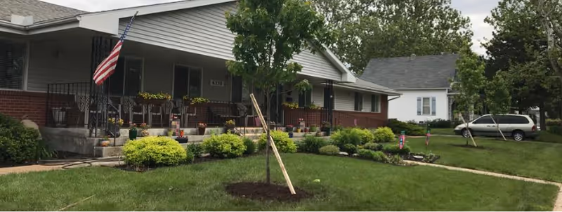 Front exterior of a single-story building with a covered porch, American flag, landscaped lawn, shrubs, and a young tree.