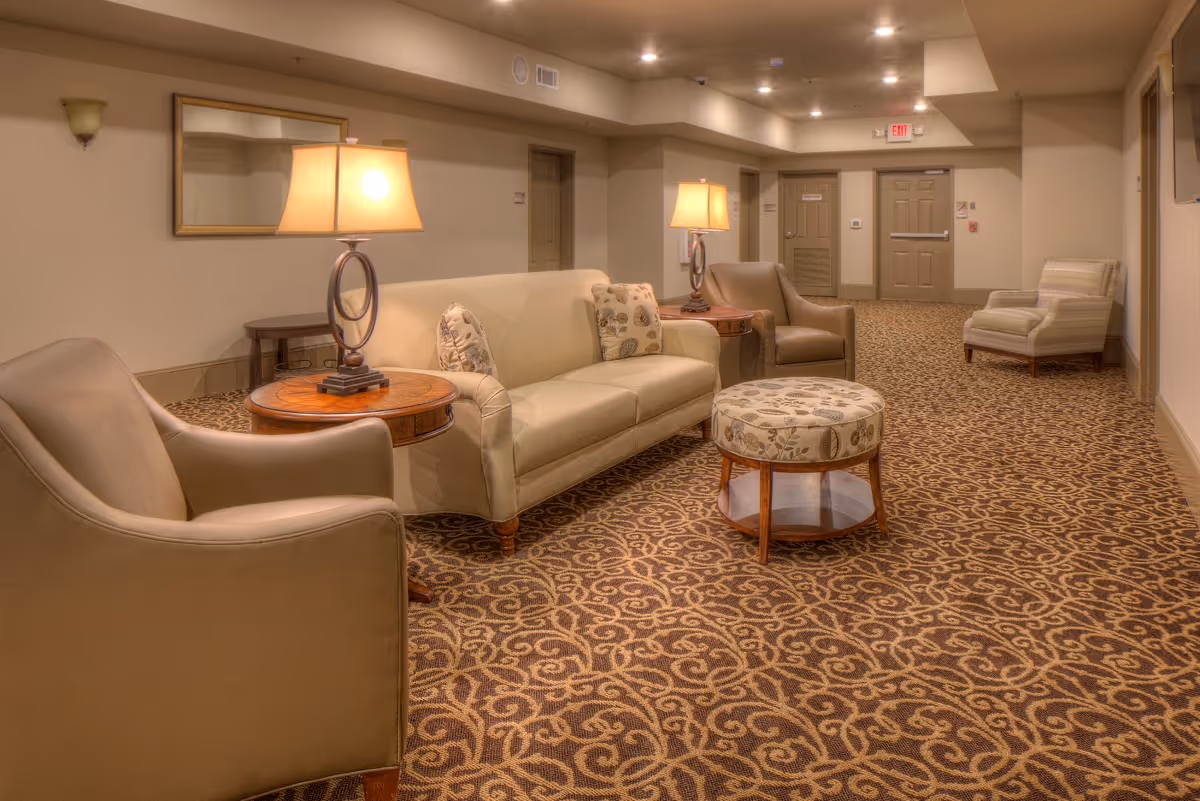 A cozy senior living facility common area with beige walls and patterned carpet. The room features a beige sofa with decorative pillows, two armchairs, a round ottoman with floral upholstery, and two wooden side tables each with a lamp. There are doors and an exit sign visible in the background.