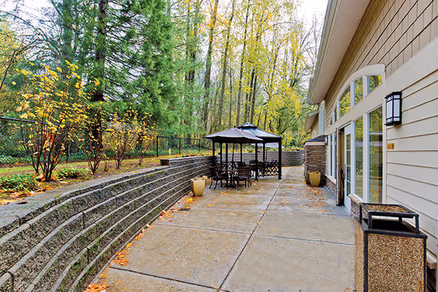 Outdoor patio area with a concrete walkway, a black metal table and chairs under a large umbrella, surrounded by a retaining wall and trees with autumn foliage. The side of a beige building with large windows and a wall-mounted light fixture is visible on the right.