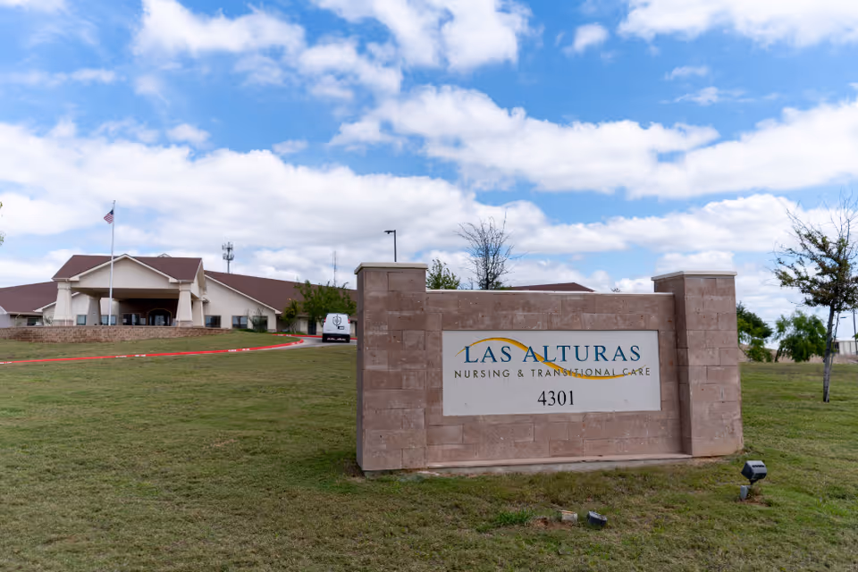 Exterior view of Las Alturas Nursing & Transitional Care facility with a large stone sign displaying the facility name and address 4301, a grassy lawn, a flagpole with an American flag, and a partly cloudy sky.