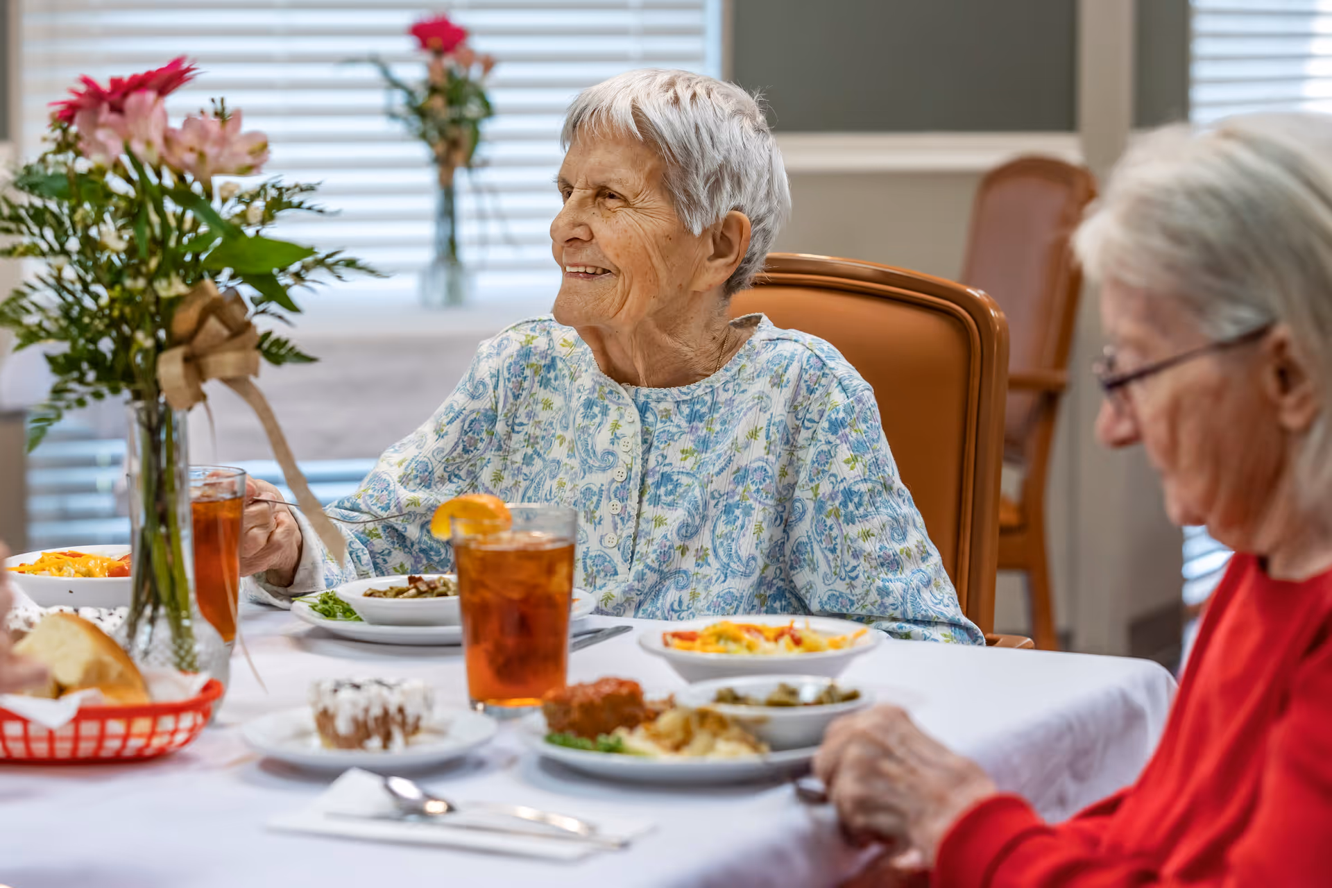 Two elderly women sitting at a dining table in a senior living facility, enjoying a meal together. The table is set with plates of food, glasses of iced tea with lemon, and a vase with flowers. One woman is smiling and looking to the side, while the other is focused on her food.
