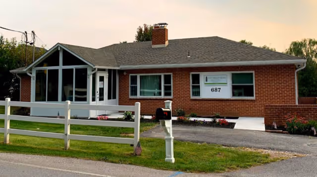 Single-story brick building with a chimney and large windows, surrounded by a white fence and green lawn. A mailbox is visible near the road, and a sign on the building reads Pine Manor Home with the number 687. Trees are in the background under a cloudy sky.