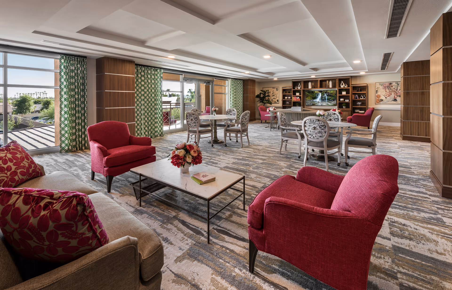 Spacious senior living common room with red armchairs, sofas, dining tables, built-in shelving and large windows.