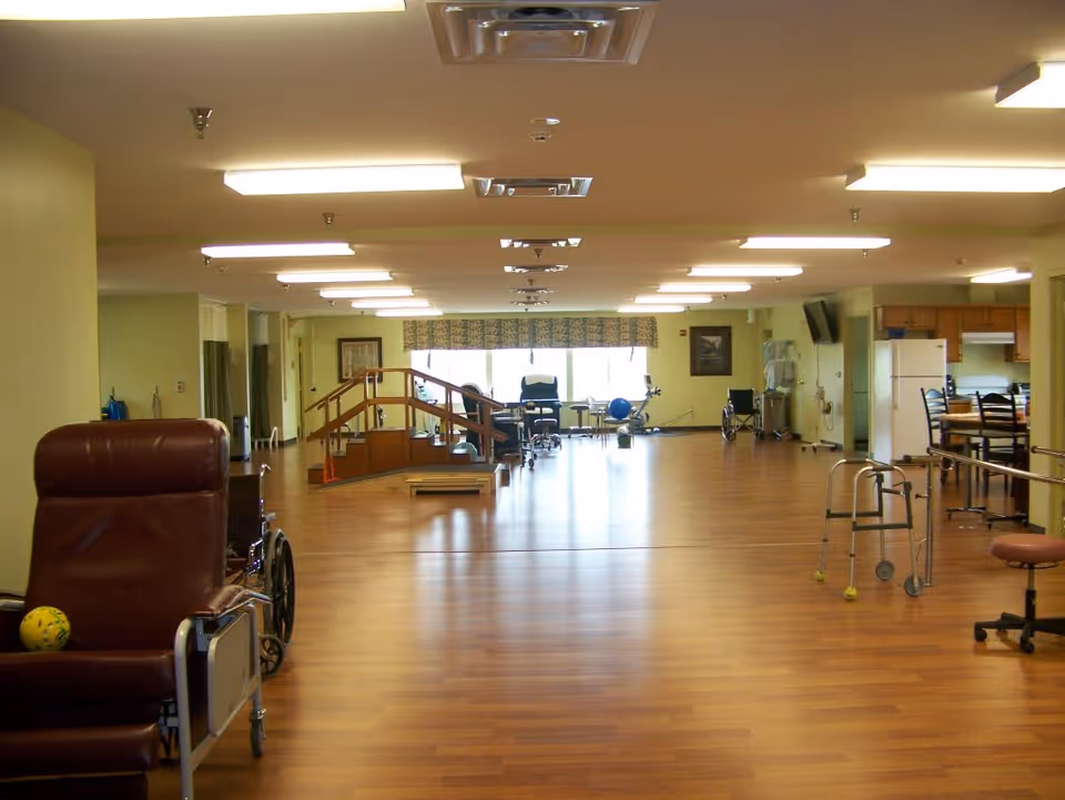 Spacious nursing center activity/rehabilitation room with wood floors, chairs, walkers and exercise equipment centered around a small ramp and open area.