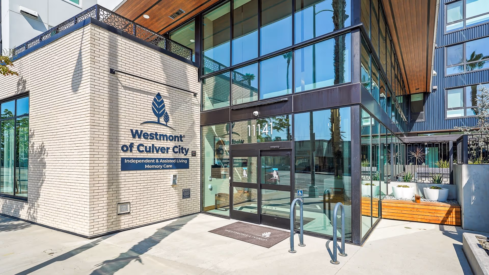 Exterior entrance of Westmont of Culver City facility with large glass windows and doors, beige brick walls, and signage indicating Independent & Assisted Living and Memory Care services.
