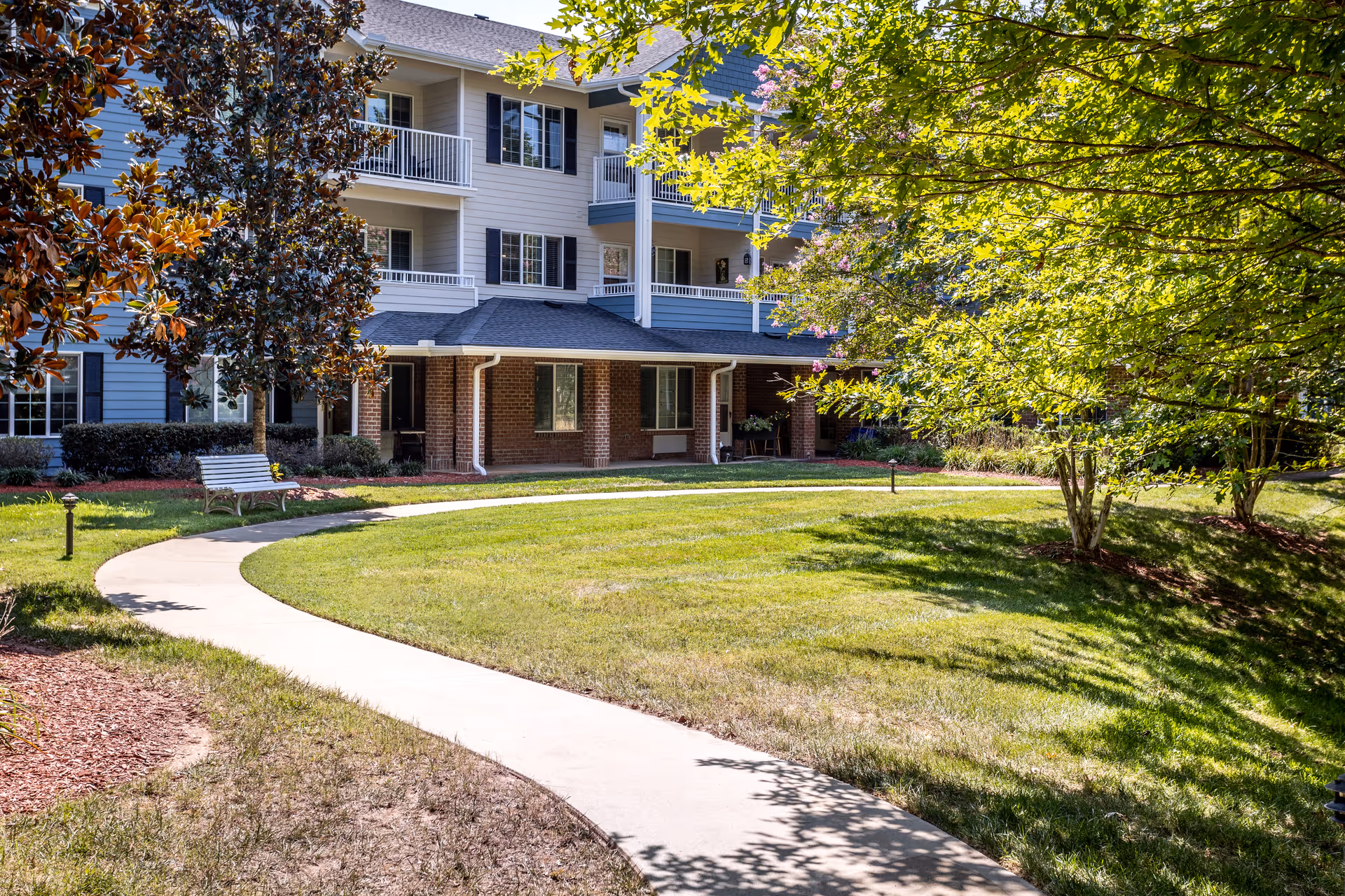 A sunny outdoor garden area at Holiday Cedar Ridge with a curved concrete walkway, green grass, trees, and a bench. The background shows a multi-story residential building with balconies and windows.