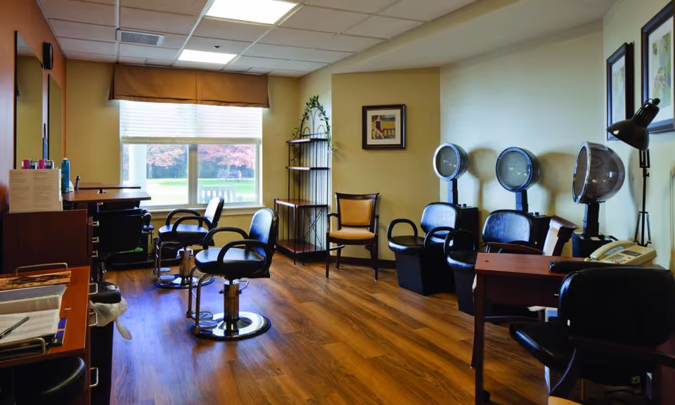 Interior view of a salon area in a senior living facility with several black salon chairs, hair drying stations, a window with a brown valance, wooden flooring, and framed artwork on the walls.