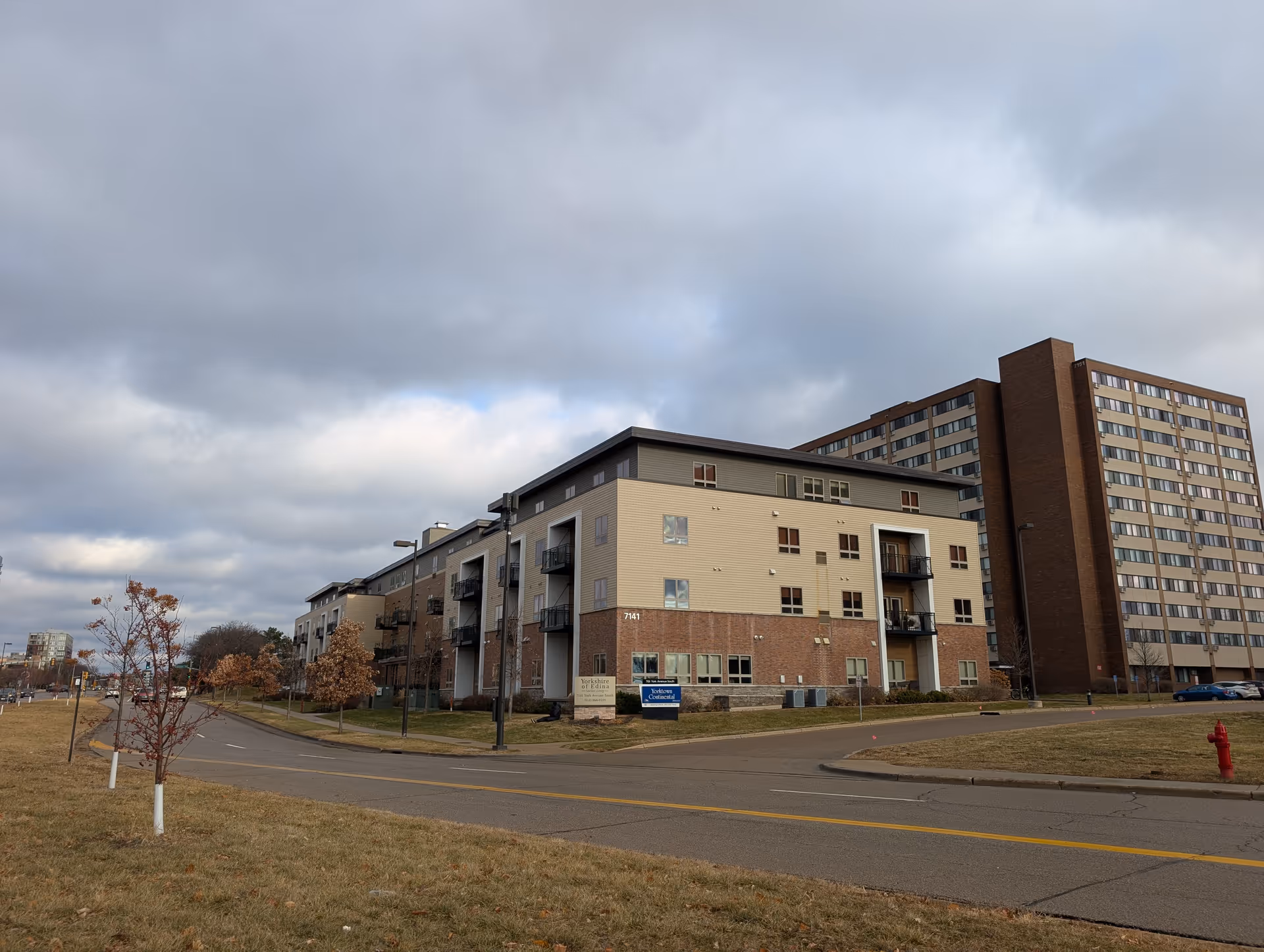 Exterior view of Yorkshire Of Edina Senior Living facility, a multi-story building with beige and brick facade, balconies, and a sign in front. The sky is cloudy and the surrounding area has a road, grass, and small trees.