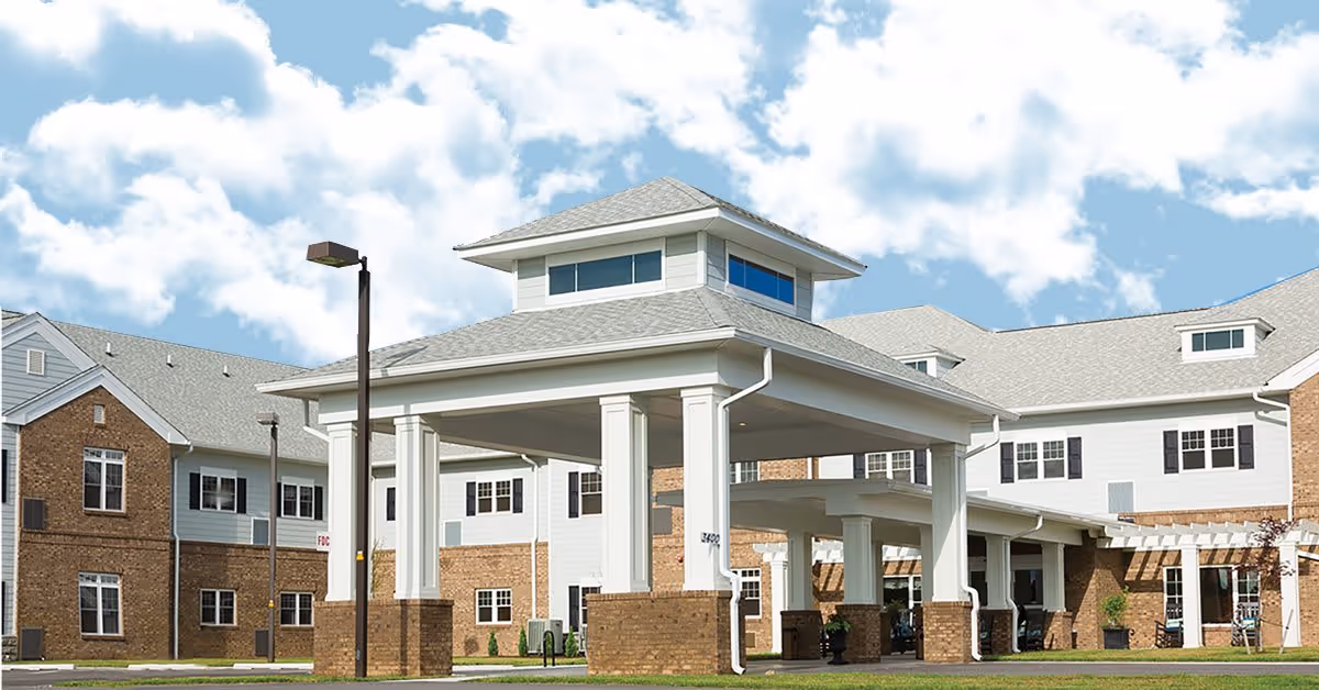 Exterior view of a senior living facility building with a covered entrance supported by white columns and brick bases. The building has multiple windows and a gray roof under a partly cloudy sky.