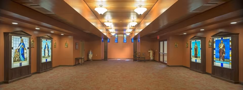 Long warm-lit interior corridor with stained-glass religious panels along both walls and a wood-paneled ceiling.