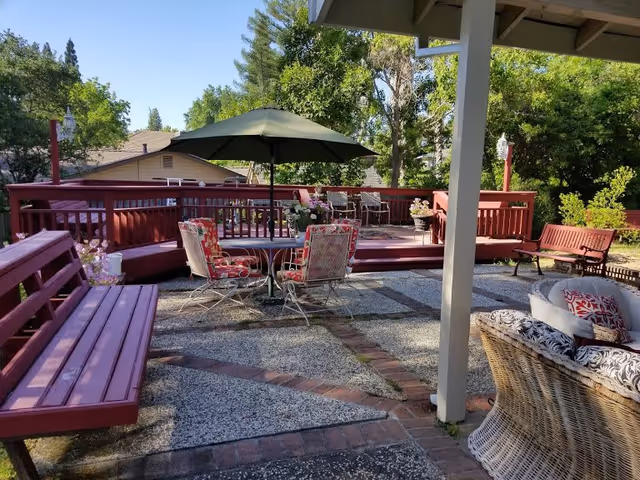 Outdoor patio area with red wooden benches, a round table with four cushioned chairs under a green umbrella, and a wicker sofa with patterned cushions. The patio is surrounded by trees and greenery under a clear blue sky.