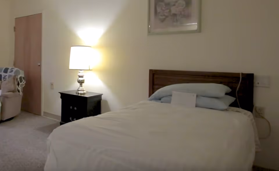 A bedroom with a neatly made bed featuring light blue pillows and a white comforter. To the left of the bed is a dark wooden nightstand with a lit table lamp. In the background, there is a closed wooden door and a beige armchair with a patterned blanket draped over it. The walls are light-colored and there is a framed picture hanging above the bed.