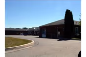 Exterior view of a single-story brick building with a paved driveway and a tall tree near the entrance under a clear blue sky.
