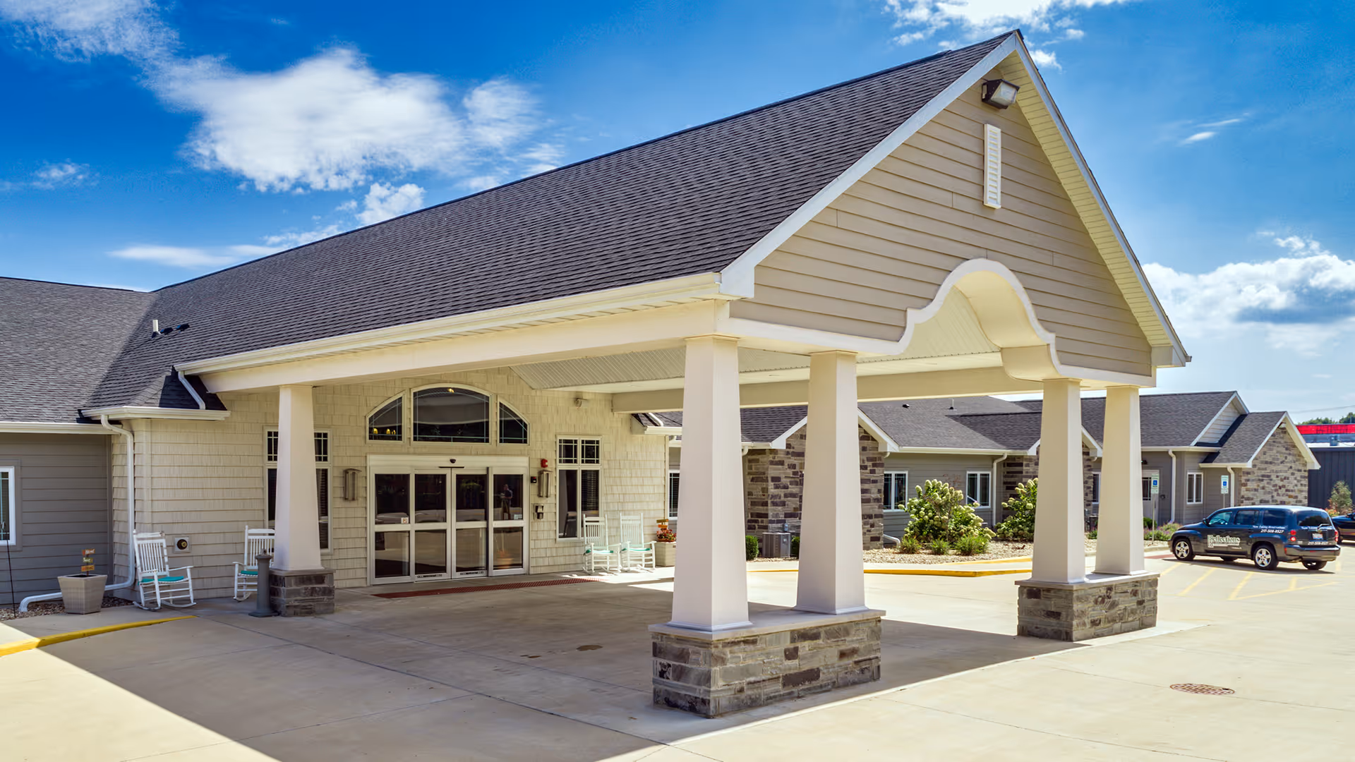 Entrance of Reflections Memory Care facility in Chatham, IL, showing a covered drop-off area with four white columns on stone bases, a paved driveway, and a building with beige siding and stone accents under a blue sky with some clouds.