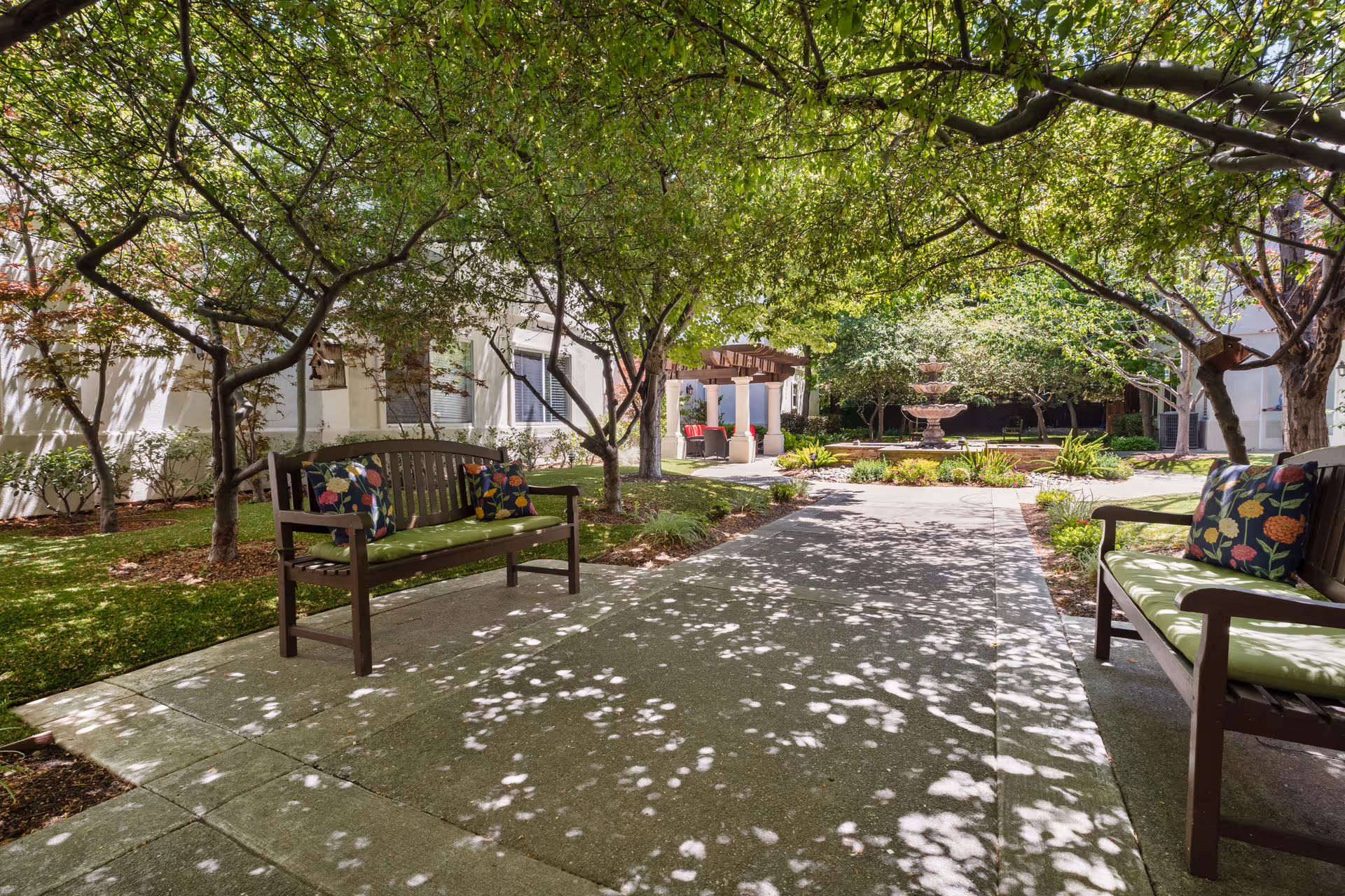 Shaded outdoor courtyard with wooden benches, leafy trees, a central fountain and a pergola seating area.