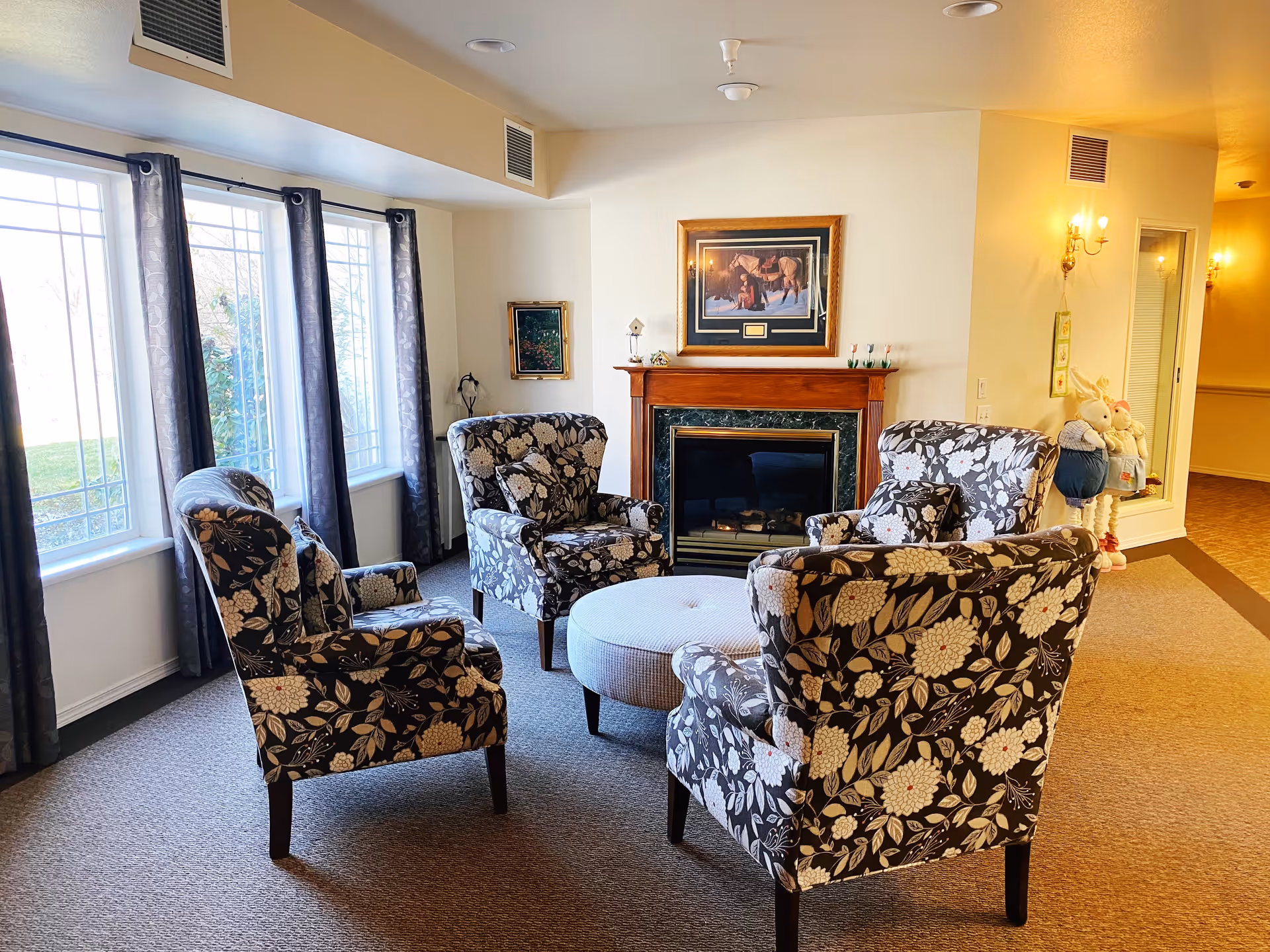 A cozy sitting area in a retirement and assisted living facility featuring four floral-patterned armchairs arranged around a round upholstered ottoman. Behind the chairs is a fireplace with a wooden mantel and a framed picture above it. Large windows with dark curtains allow natural light to fill the room. The walls are light-colored, and there are decorative items including two stuffed bunny dolls near a mirror on the right side.