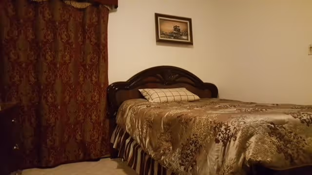 A bedroom with a decorative wooden headboard, patterned bedspread, matching drapes, and a small framed picture on the wall.