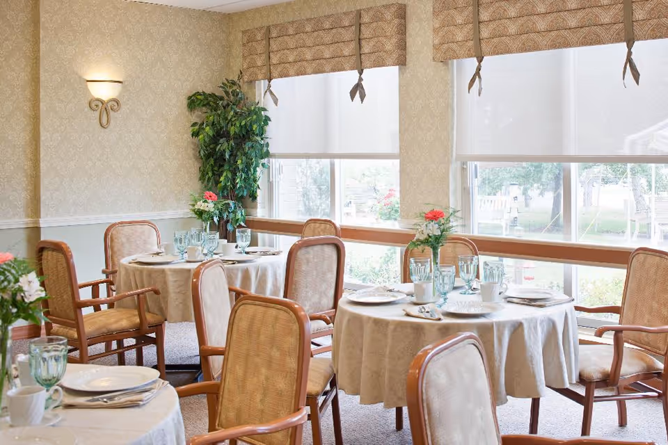 Sunlit dining room with round tables set with tablecloths, glassware, and chairs beside large windows and plants.