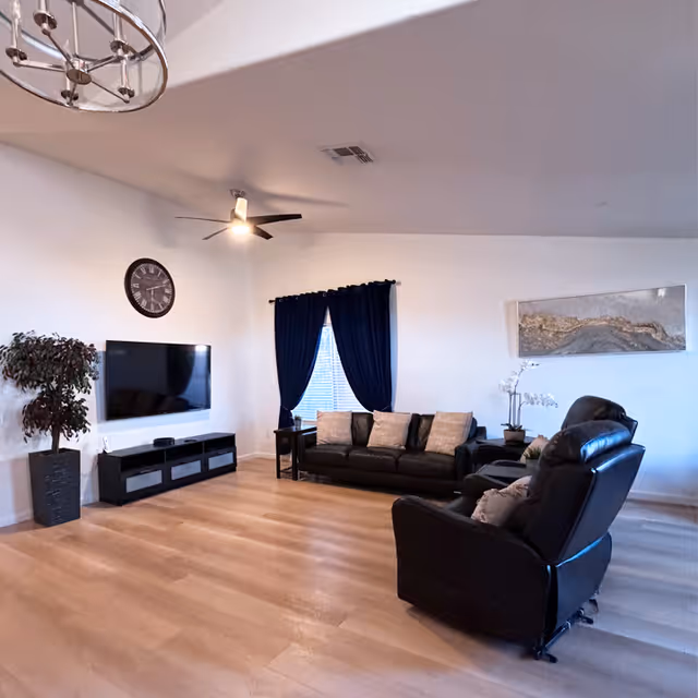 A spacious living room with light wooden flooring, a black leather sofa and recliner, a wall-mounted flat screen TV, a large clock on the wall, a potted plant, dark blue curtains covering a window, and a modern ceiling fan with light.