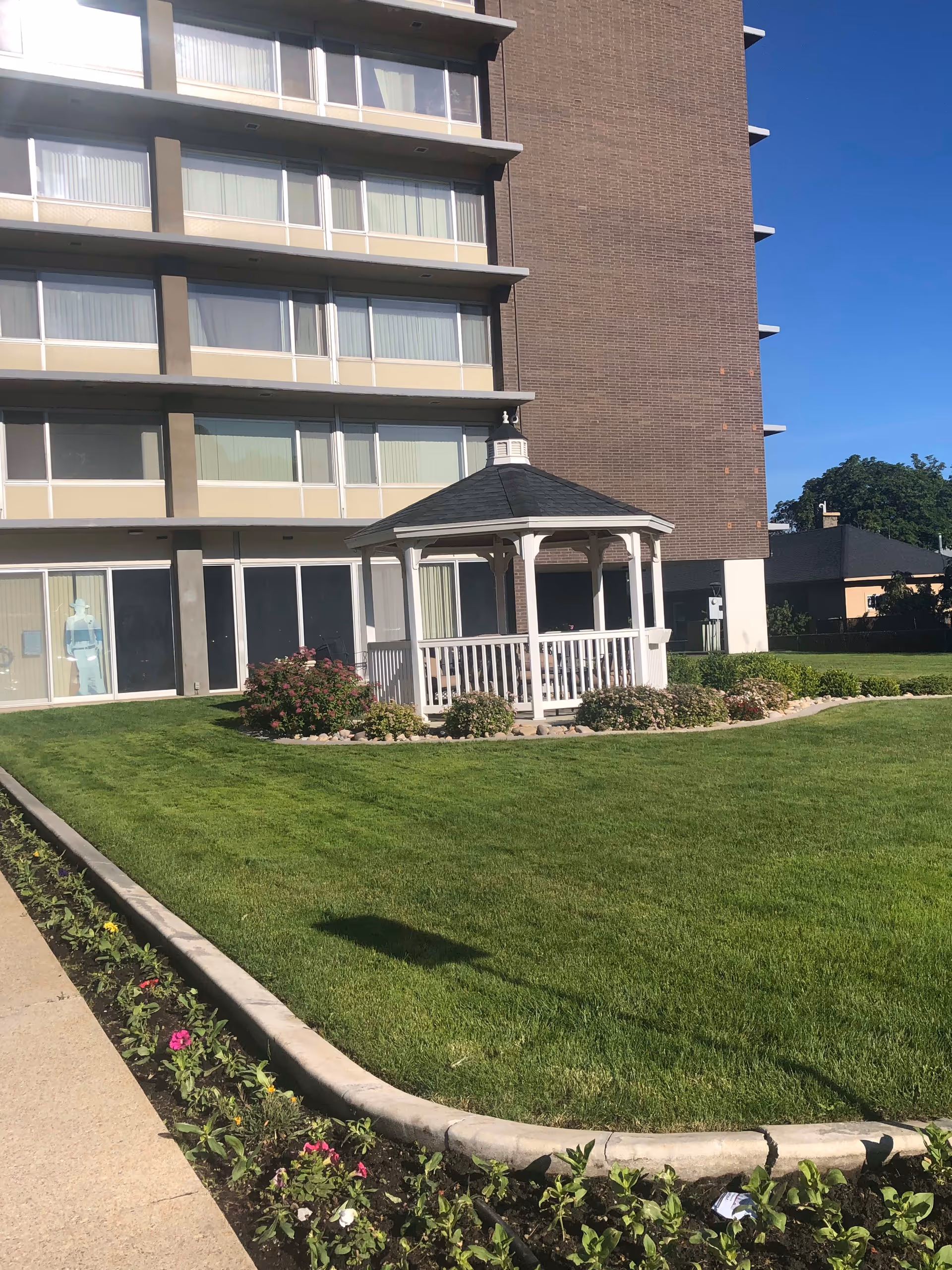 Multi-story building facade with a white gazebo on a manicured lawn and flowerbeds in front.