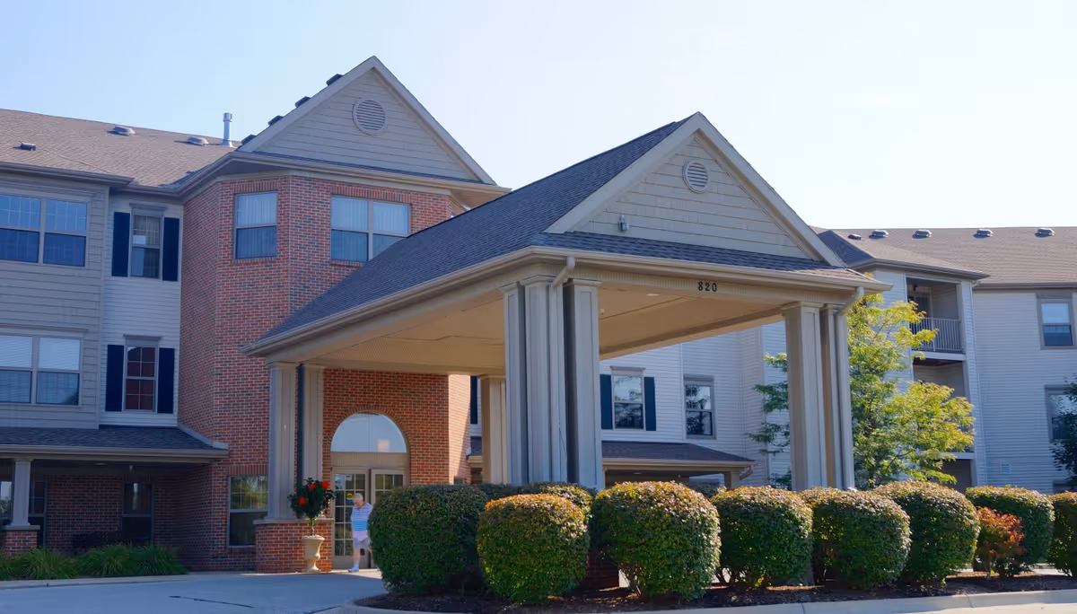 Exterior view of a senior living facility building with a covered entrance supported by columns. The building has a combination of brick and siding with multiple windows and a well-maintained hedge in front. A person is visible near the entrance.