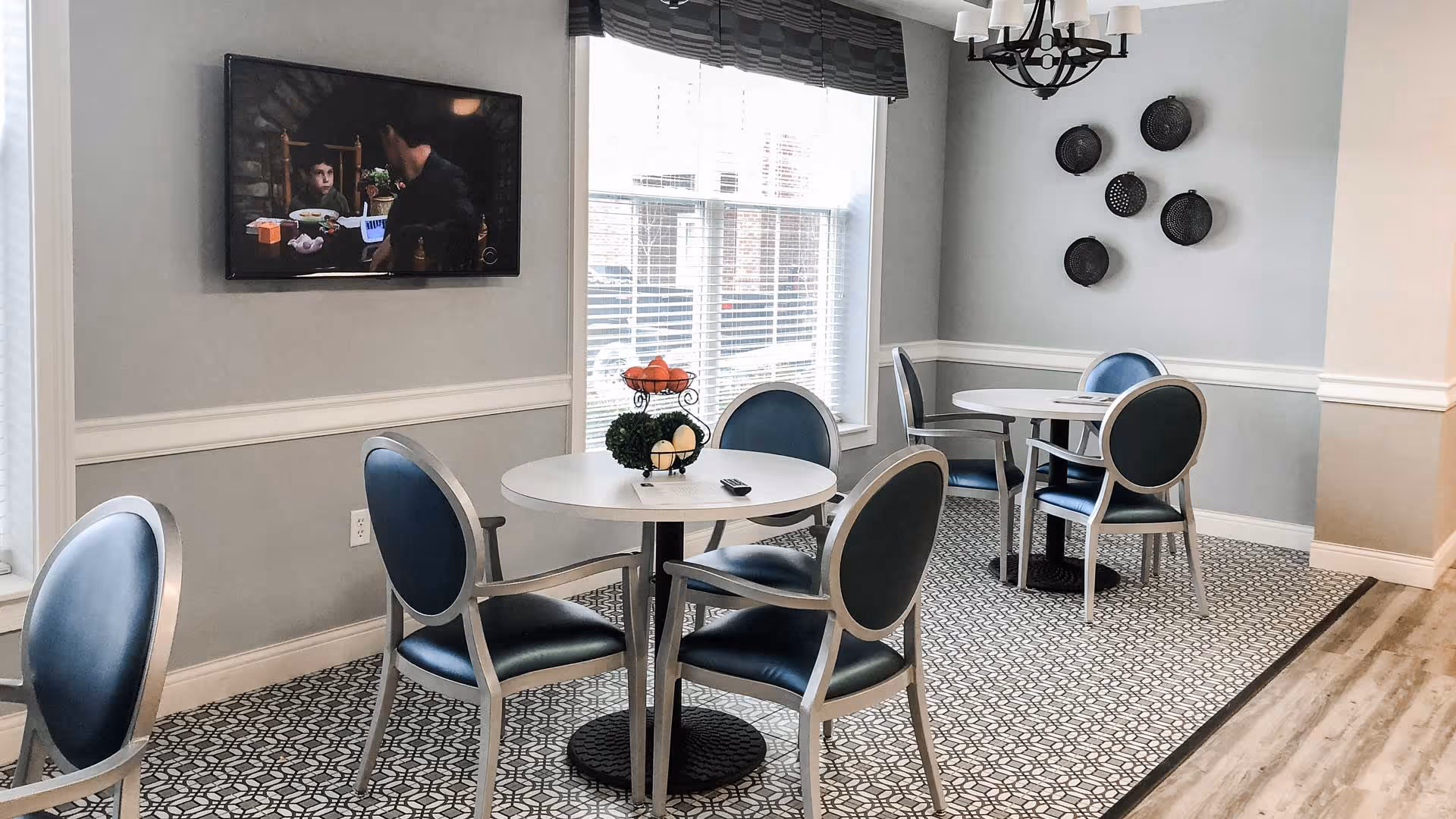 A dining area with two round white tables, each surrounded by four blue cushioned chairs with silver frames. A flat-screen TV is mounted on the wall showing a scene from a show. There is a large window with blinds letting in natural light. The walls are painted light gray with white trim, and decorative black baskets are hung on one wall. The floor has a patterned carpet bordered by wood flooring.