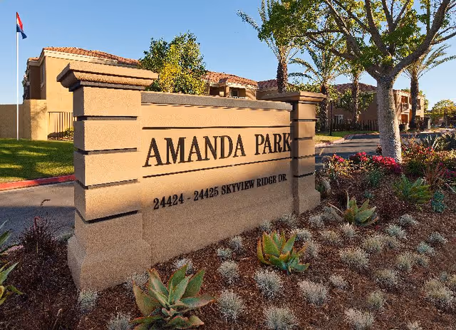 Stone sign at the entrance of Amanda Park facility with the address 24424 - 24425 Skyview Ridge Dr., surrounded by landscaped plants and trees, with buildings and a flagpole visible in the background under a clear blue sky.