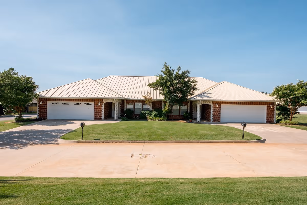 Front exterior view of a single-story brick building with a light-colored metal roof, two garages, a driveway, and a well-maintained lawn with trees on either side under a clear blue sky.