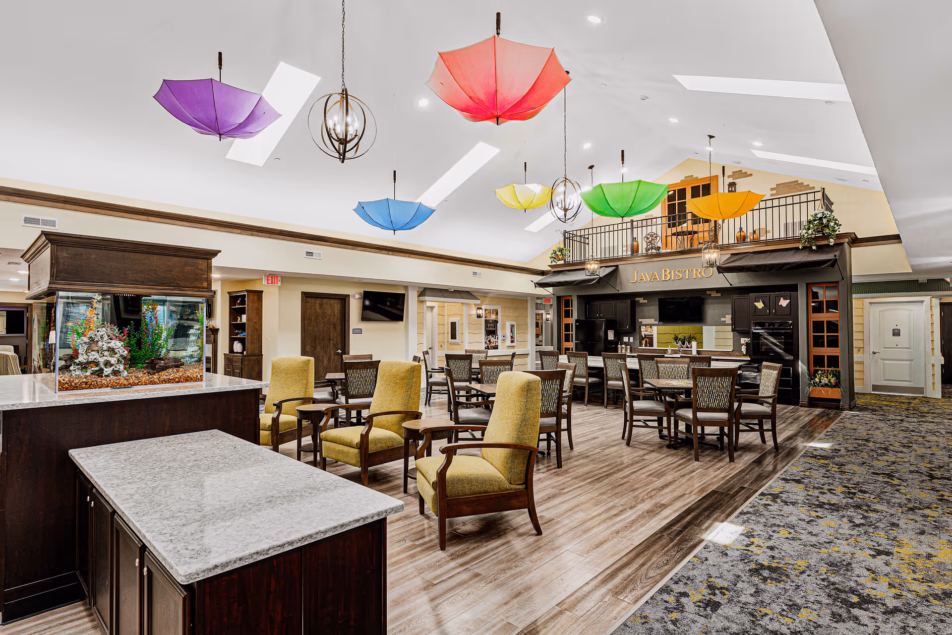 A bright and spacious dining area in an assisted living facility featuring several wooden tables and chairs, cushioned armchairs, a large aquarium on a dark wood cabinet, and a coffee bar labeled 'Java Bistro'. The ceiling has colorful umbrella decorations hanging from it, and the floor is a combination of wood and patterned carpet. Skylights and modern pendant lights provide ample lighting.