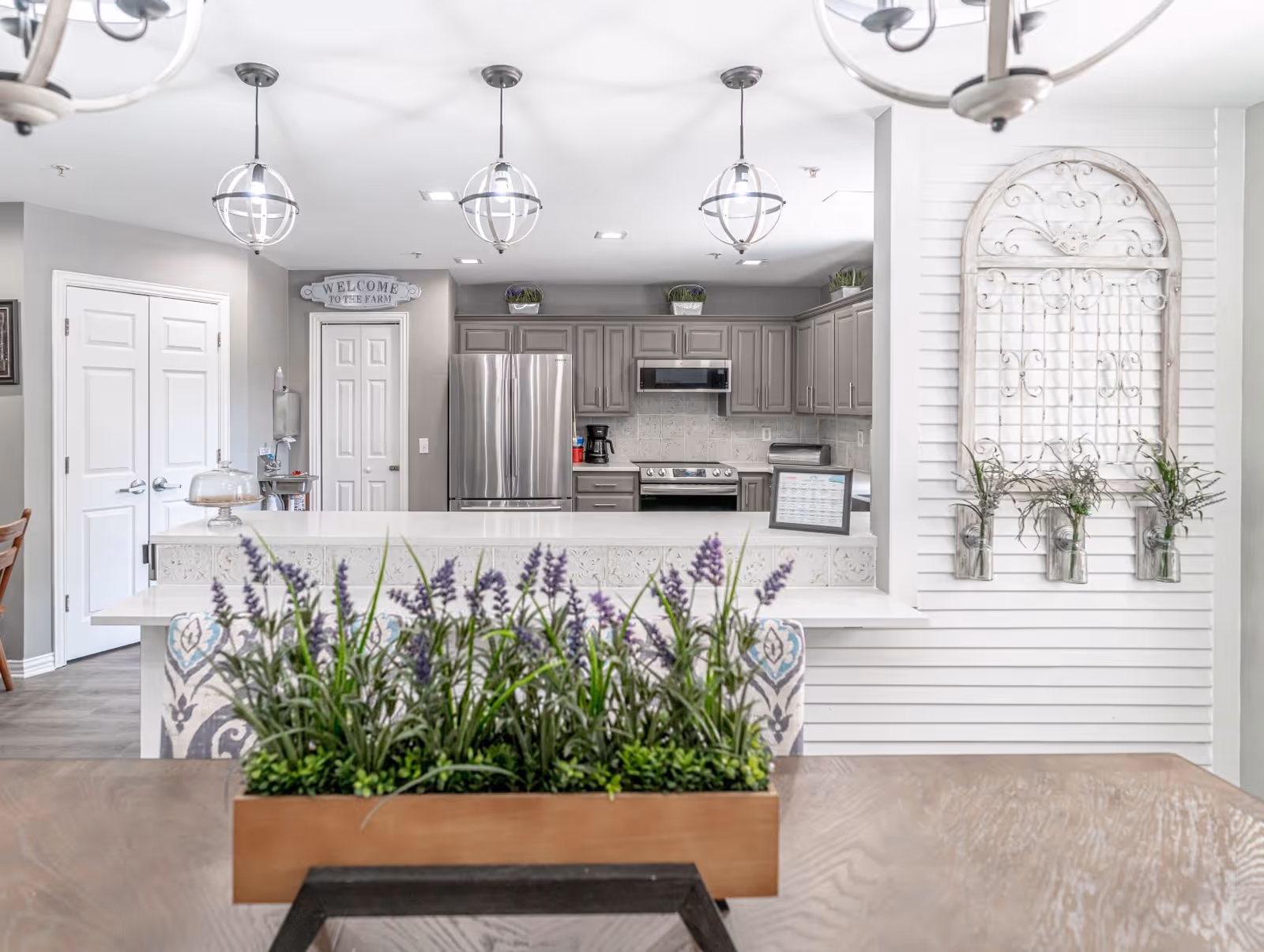 Bright open kitchen with a large island, stainless steel refrigerator, hanging globe pendant lights, and a table planter in the foreground.