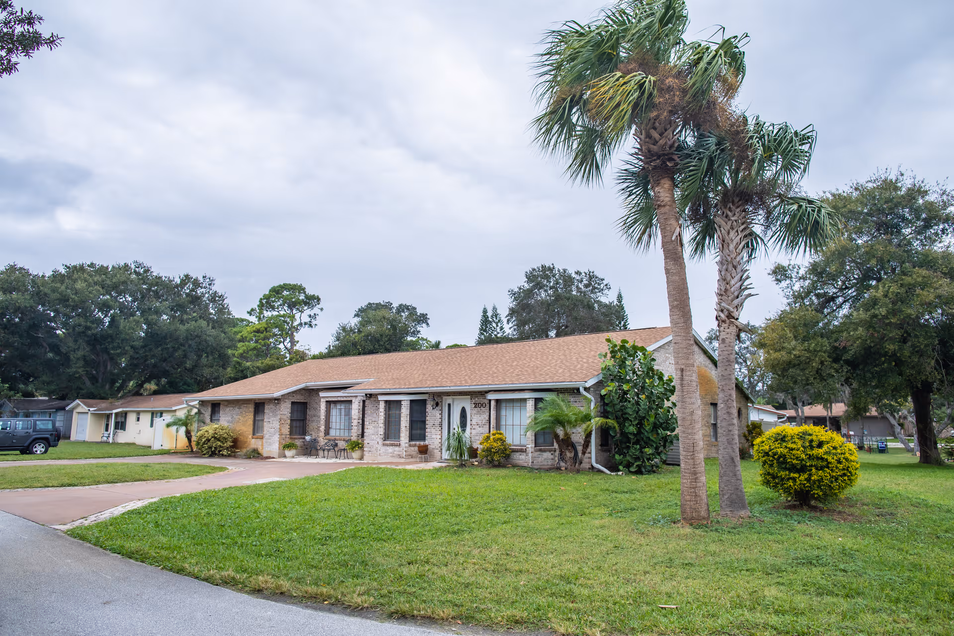 Single-story brick building with a brown shingle roof surrounded by green grass, palm trees, and other shrubs under a cloudy sky. There is a driveway leading up to the building and a parked vehicle to the left.