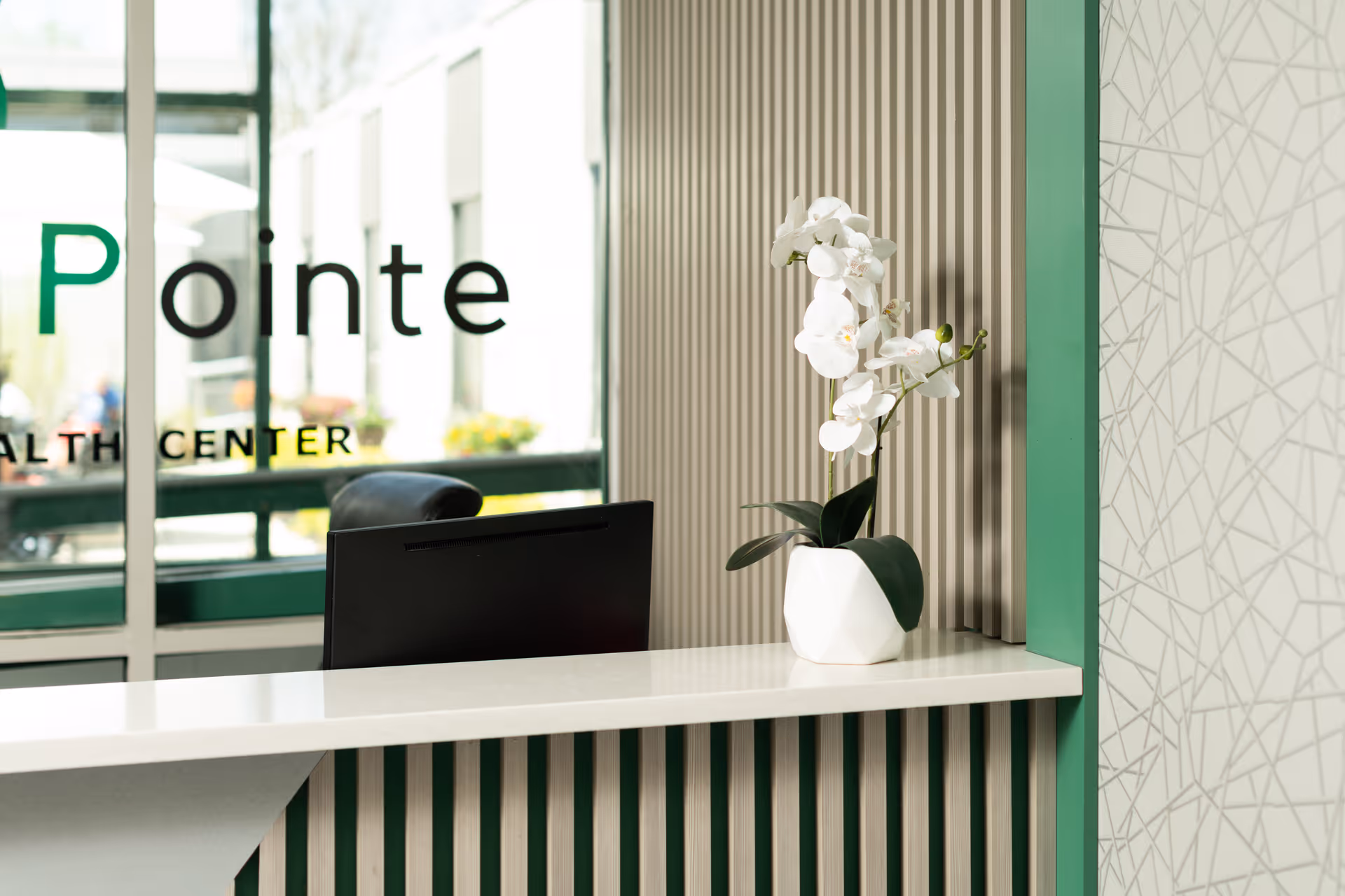 Reception desk area with a white countertop, a black computer monitor, and a white vase holding white orchids. Behind the desk is a window with partial text reading 'Pointe' and 'Health Center'. The wall has vertical wooden slats and a green frame with a patterned wallpaper on the right side.