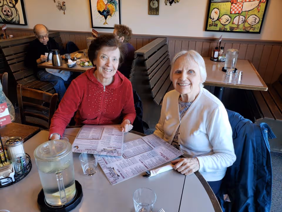 Two elderly women sitting at a restaurant table holding menus and smiling at the camera. In the background, two other elderly people are seated at another table eating. The walls have colorful paintings of farm animals.