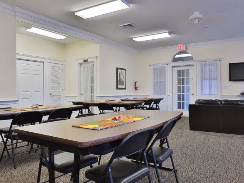 A well-lit common area with several rectangular tables and black folding chairs arranged around them. The tables have colorful placemats and small decorative centerpieces. In the background, there is a brown leather couch, a wall-mounted TV, and a door with an exit sign above it. The walls are light-colored with white trim, and there is a framed picture and a fire extinguisher on one wall.