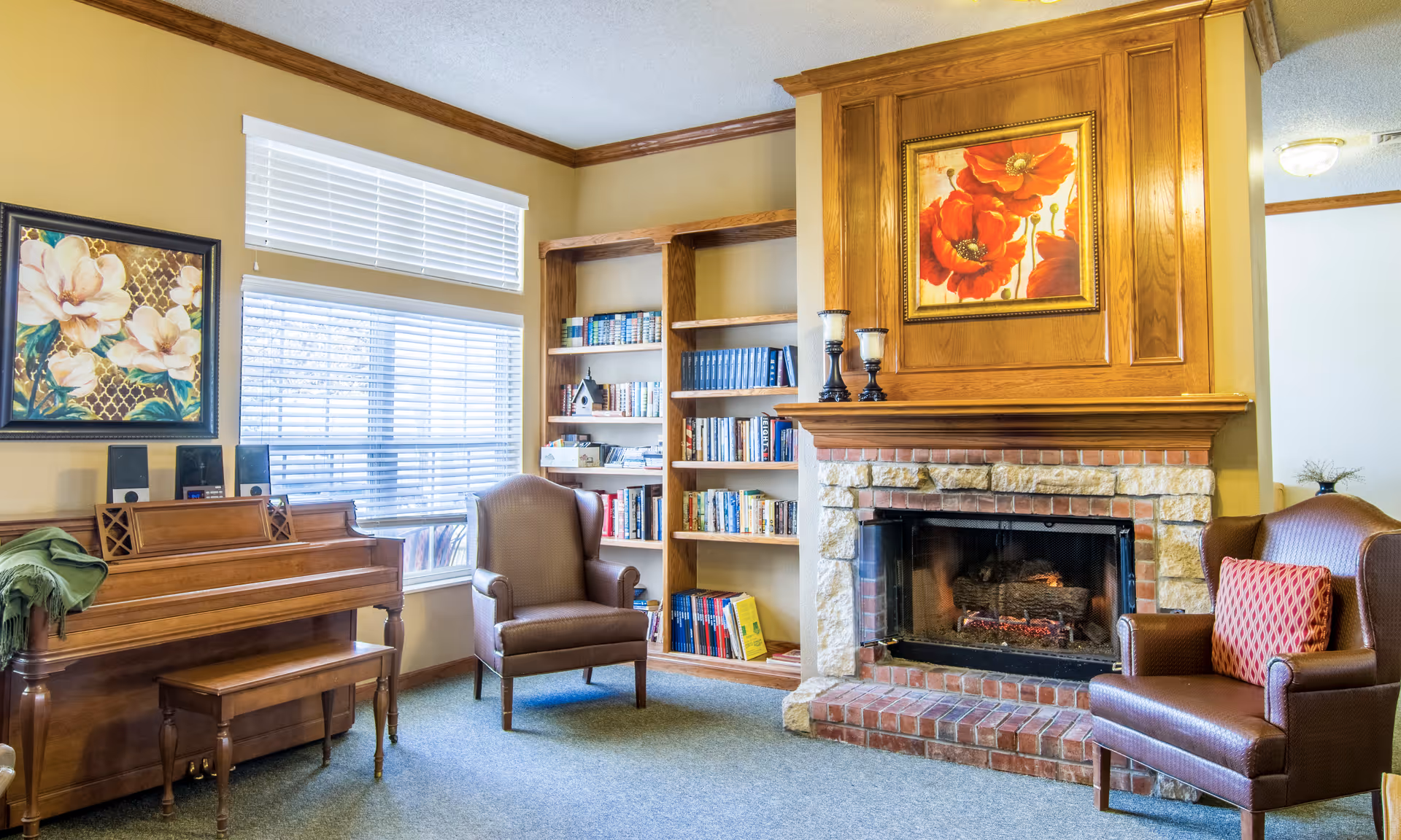 A cozy living room area featuring a wooden piano with a bench, two brown upholstered armchairs, a stone and brick fireplace with a wooden mantel, and built-in wooden bookshelves filled with books. The walls are painted beige with wooden crown molding, and there are two windows with white blinds letting in natural light. Above the fireplace hangs a framed painting of red flowers, and another floral painting is on the wall above the piano.