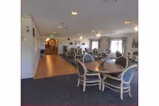 Dining area with several round tables and upholstered chairs, carpeted floor and windows letting in natural light.
