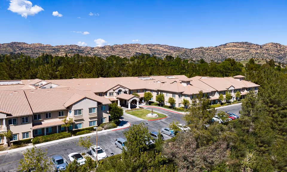 Aerial view of the Fairwinds - West Hills senior living building with a circular driveway, parked cars, surrounding trees and distant hills under a blue sky.
